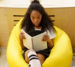 Young Black girl reading a book while sitting in a bright yellow beanbag chair