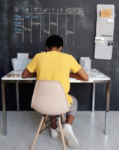 Black student in a yellow shirt facing the wall and working at a white desk in front of a black chalk wall with a weekly schedule on it