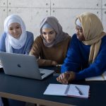 Three young women wearing hijab working at a table with books and a laptop